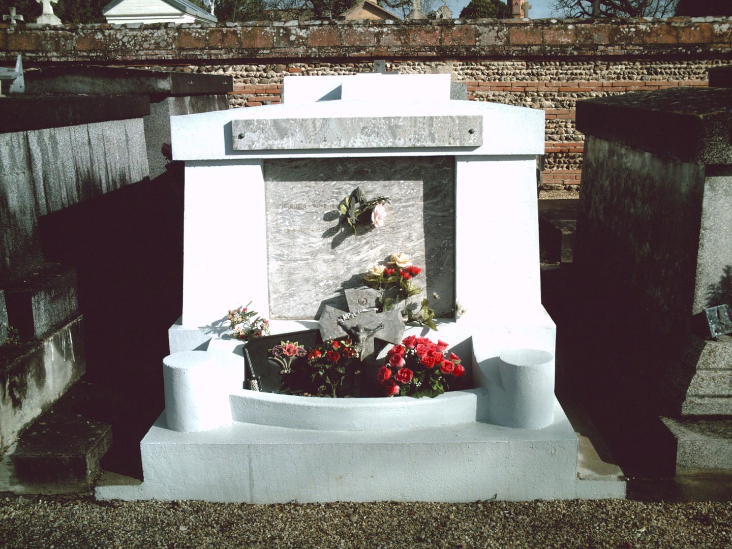 Tombe en marbre blanc dans un cimetière, ornée de roses rouges et blanches devant une plaque de marbre gris et un mur de briques en arrière-plan.