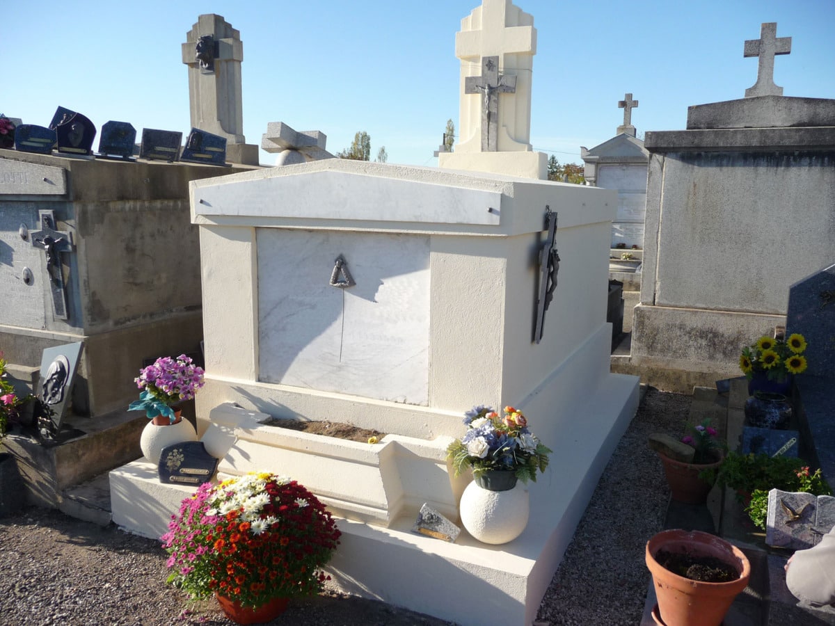Tombeau familial blanc au premier plan dans un cimetière, décoré de fleurs colorées et de crucifix. Stèles funéraires en arrière-plan.
