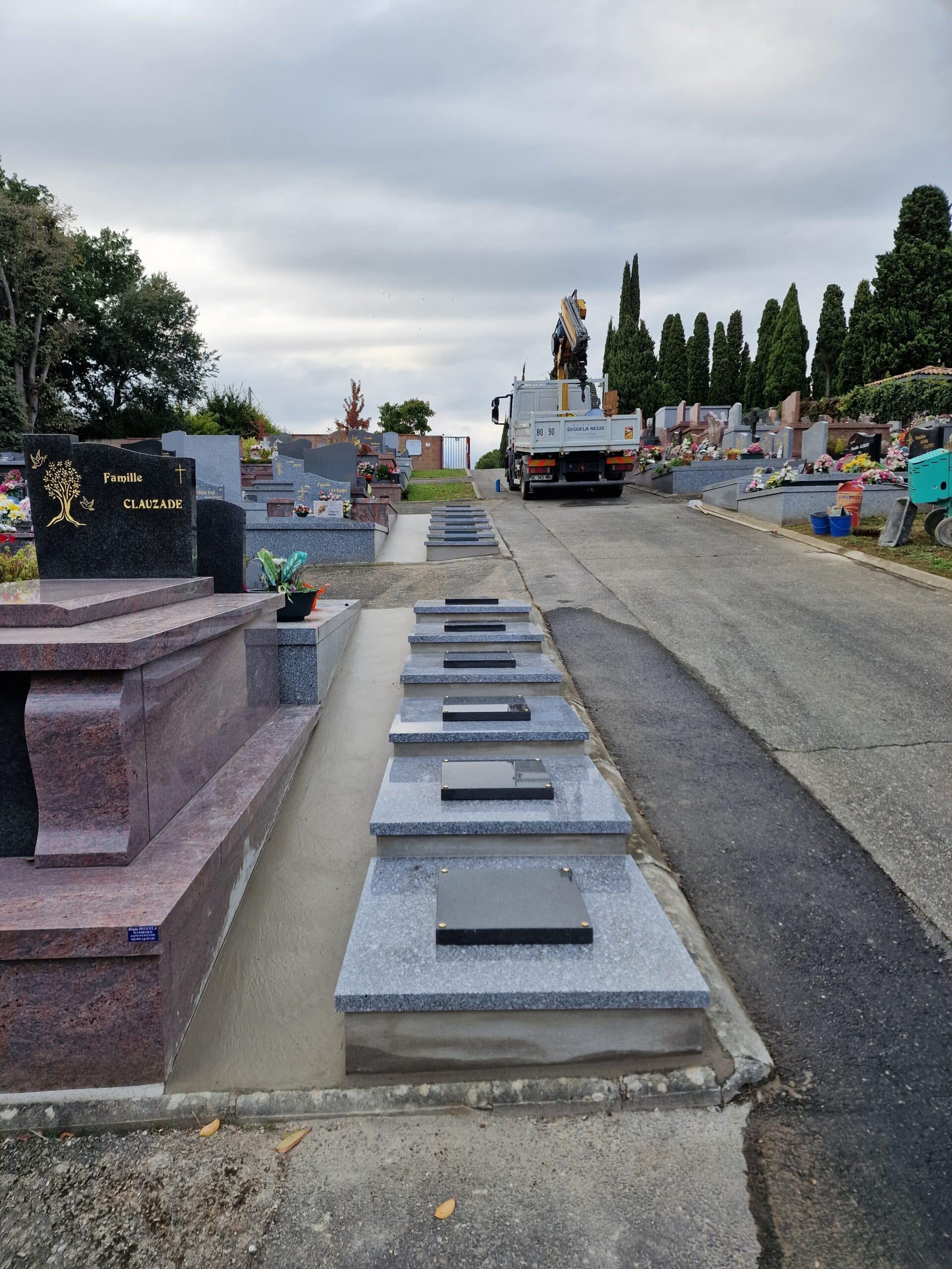 Nouveaux caveaux gris alignés dans un cimetière, tombe Clauzade au premier plan et camion grue Séguéla Régis au loin.