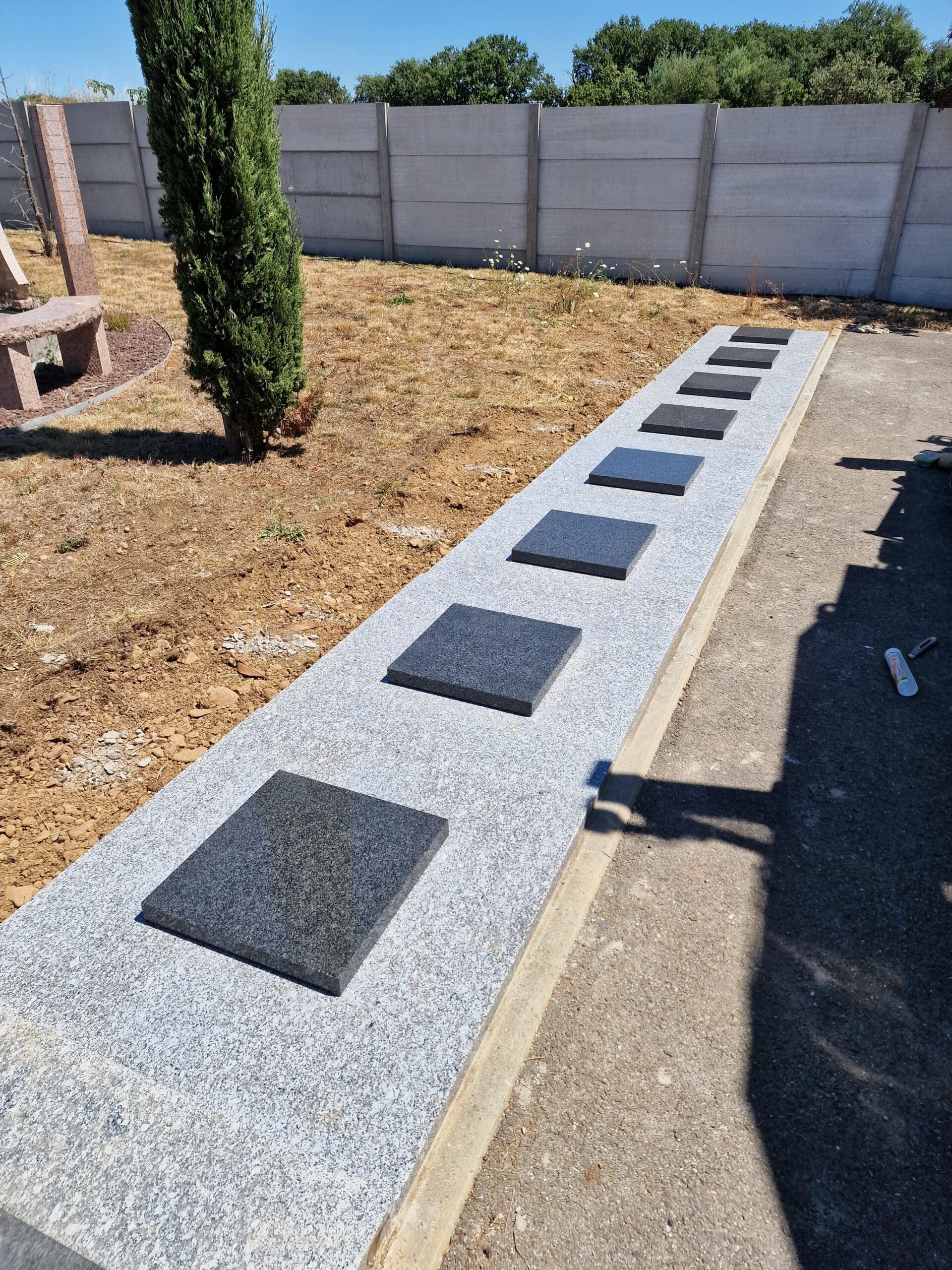 Longue dalle en granit gris surmontée de carreaux noirs polis dans un espace extérieur avec cyprès et clôture.