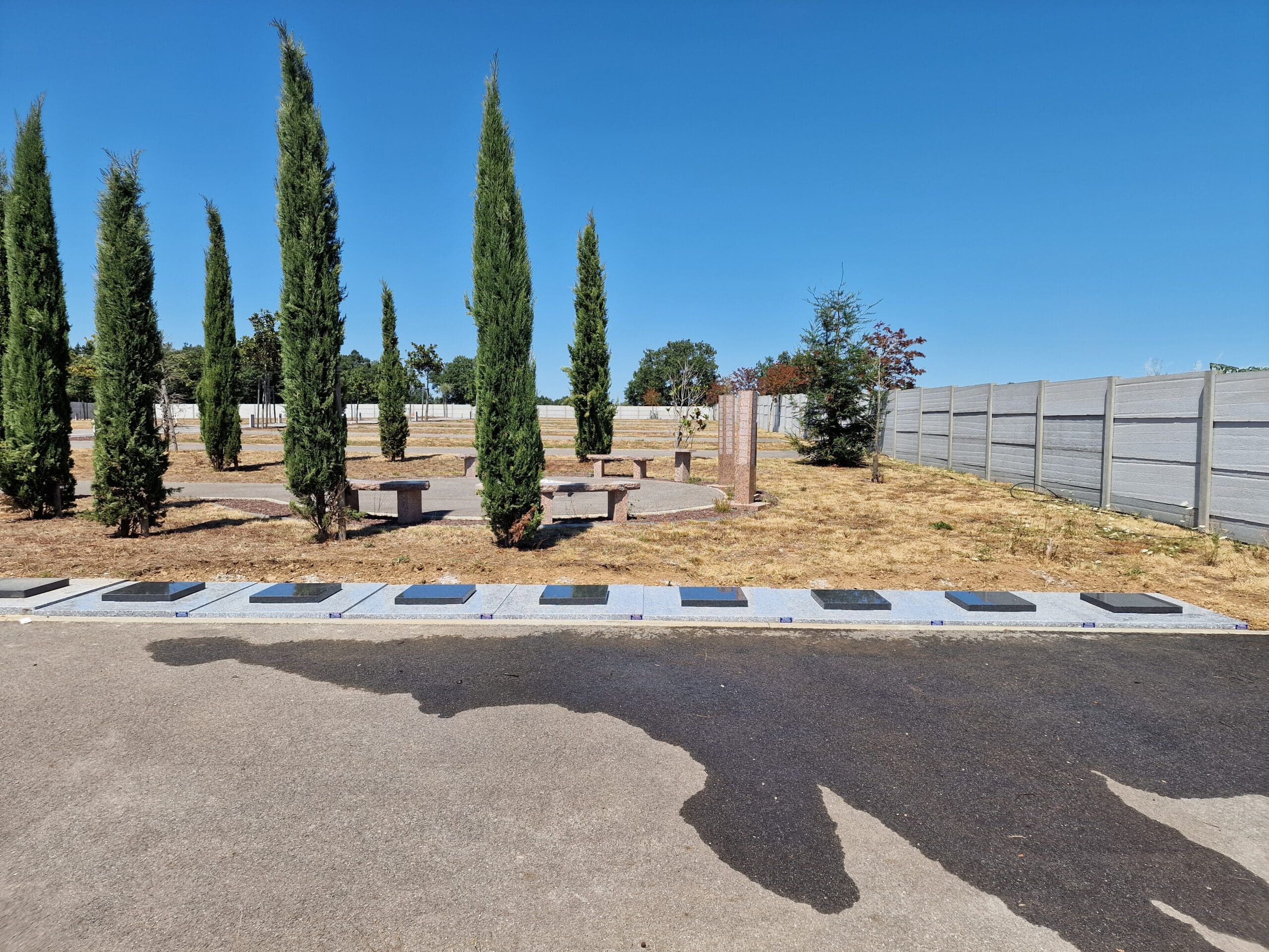 Jardin commémoratif avec cyprès, bancs en pierre et plaques funéraires alignées sous un ciel bleu dégagé.
